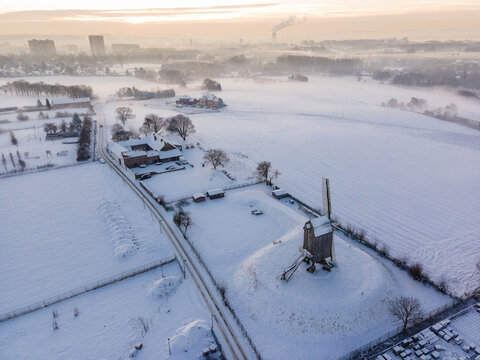 Aerial shot of a white winter landscape with the Luizenmolen windmill in the foreground, and the town of Anderlecht and Brussels skyline visible in the background under soft morning light
