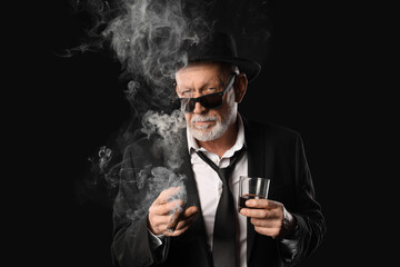 Old businessman with glass of whiskey smoking cigar on black background