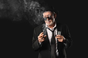 Old businessman with glass of whiskey smoking cigar on black background