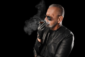Stylish old man in leather jacket smoking cigar on black background