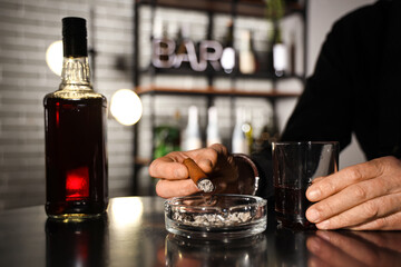 Stylish old man with glass of whiskey putting out his cigar in ashtray at table in bar, closeup