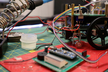 Photo of process of creating mini robot. On background of blurring parts, soldering iron, tools, microcircuits, workspace. Robot is not finished, it has eyes, wheels, microcircuits. Hobby, craft, DIY.