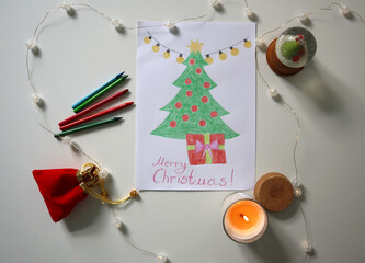 Flat lay of hand drawn Christmas tree with gift and Merry Christmas text, surrounded by crayons, candle, snow globe, and fairy lights. Childrens Christmas card. Festive content, kids projects, cards.