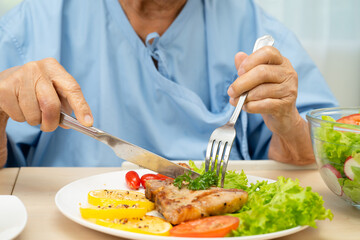 Asian senior woman patient eating pork chop stake and vegetable salad for healthy food in hospital.