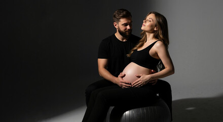 Pregnant woman with partner in black outfits touching belly on exercise ball. Active maternity lifestyle for prenatal fitness and wellness program documentation