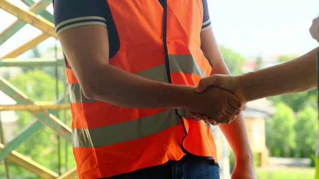 Worker in orange safety vest shakes hands with taskmaster on construction site closeup. Young man gets job at building project in city