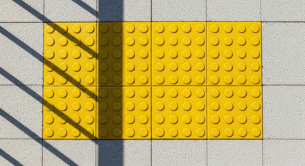 Yellow tactile paving blocks arranged on a concrete surface, casting shadows from nearby structures, enhancing urban accessibility and safety for pedestrians navigating the area