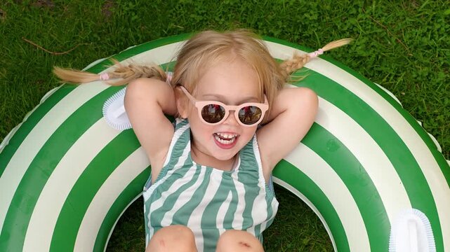 A smiling little child girl in a swimsuit lies on a swimming circle on the grass and enjoys summer and soapy puddles, the concept of happy summer. View from above