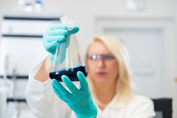 Blonde woman in rubber gloves inspecting a colored solution in a flask while standing in a biochemistry laboratory with HPLC and lab equipment