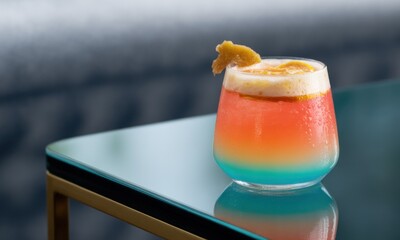 Colorful layered cocktail in a teal glass, garnished with orange zest, on a mirrored surface