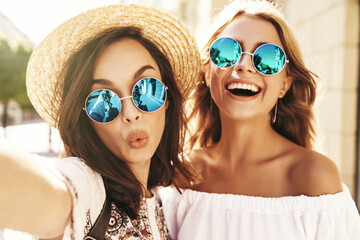 Two young female smiling hippie brunette and blond women models in summer sunny day in white hipster clothes taking selfie photos for social media on smartphone on the street background. Giving kiss