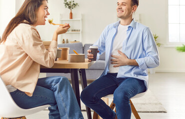 Obraz premium Content and smiling young family couple is savoring a delivered meal, eating takeout food, talking and laughing, enjoying hot beverages. They appear full and ready for a relaxed day at home.