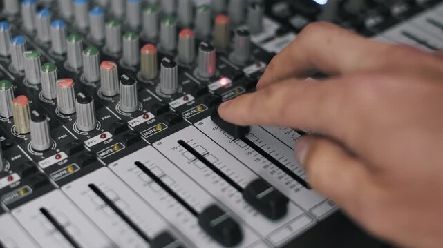 Close-up of sound engineer's hand pushing up a fader on a mixing console, adjusting volume levels on professional audio equipment in a recording studio. Colorful knobs and mute buttons are visible.