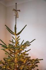 Lit Christmas Tree Topped with a Bow. Minimalist Christmas tree with white bow topper and warm string lights in modern interior