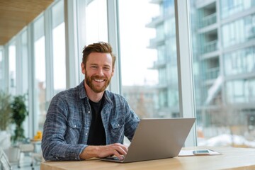 Fototapeta premium A happy man sitting at a table, using a laptop in a bright, modern home office. the man he is smiling while working online or looking for inspiration.