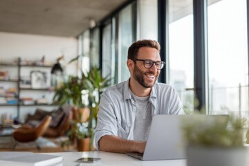 Obraz premium A happy man sitting at a table, using a laptop in a bright, modern home office. the man he is smiling while working online or looking for inspiration.