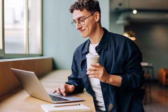 Young man working on laptop holding a coffee cup with a smile - Powered by Adobe