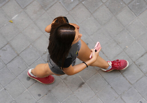 Chica joven dando paso y zancada con zapatillas rojas y pantal&oacute;n corto caminando por la calle vista desde arriba en perspectiva cenital con c&aacute;mara de dron con tel&eacute;fono m&oacute;vil en la mano