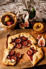 Rustic homemade fig galette on parchment paper. A dark and moody food photography still life with low key lighting, served on a vintage wooden table with a wicker basket and lace.