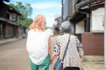 Fototapeta premium Two women walking down a street, one of them wearing a hat. The woman on the left is holding the hand of the woman on the right