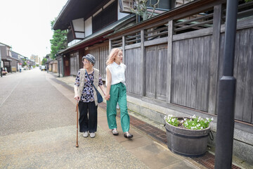 Fototapeta premium Two older women are walking down a street, one of them holding a cane. They are both wearing green pants and white shirts. The street is lined with wooden buildings, giving the scene a quaint