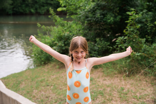Smiling girl in swimsuit with arms raised near riverside greenery.