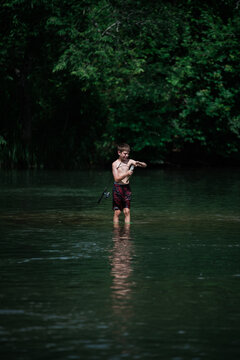 Boy fishing while standing waist-deep in calm, forested river.
