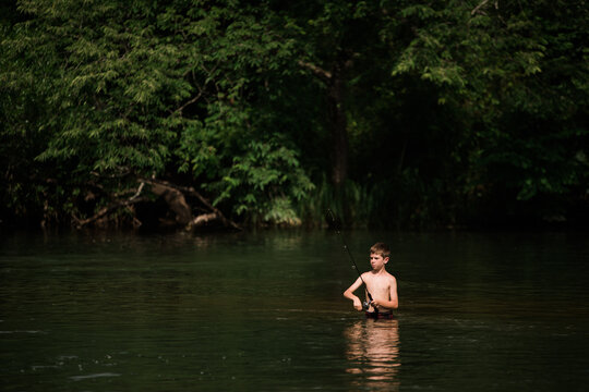 Boy fishing while waist-deep in calm river surrounded by forest.