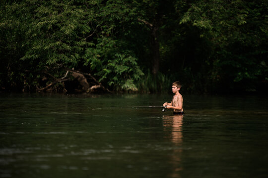 Boy fishing waist-deep in calm forest river on a sunny day