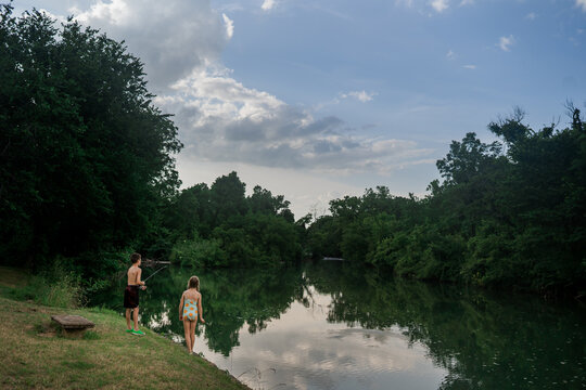 Boy fishing and girl watching beside calm river under cloudy sky.