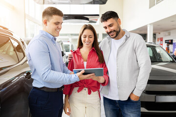 Obraz premium Smiling couple reviewing car details on tablet with dealer, happy man and woman checking purchase agreement inside showroom. Lifestyle, transport and consumer trust