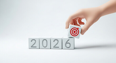 Hand placing target cube on top of blocks spelling out the year twenty twenty six on white surface