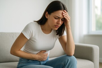 Woman experiencing stomach pain and headache while sitting on a couch in a bright room, expressing discomfort and fatigue indoors in natural light. Ai generative