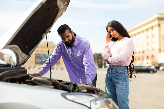 Stressed Indian couple looking at broken car engine in open hood outdoors. Woman touching head, man inspecting. Breakdown situation during travel, seeking roadside help, closeup - Powered by Adobe