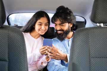 Smiling Indian couple sitting in backseat, happily browsing smartphone together. Shows joyful connection, technology use and relationship harmony, closeup
