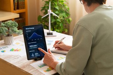 Person monitoring and planning sustainable energy consumption using a digital tablet displaying wind power performance data, surrounded by green plants and a miniature wind turbine