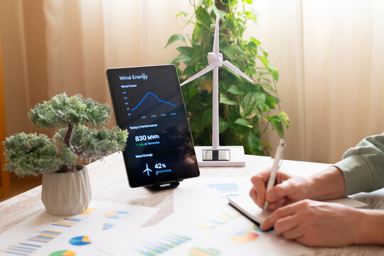 Hands using a tablet app to monitor wind power performance, taking notes beside a miniature wind turbine and potted plant, symbolizing green technology and sustainable planning