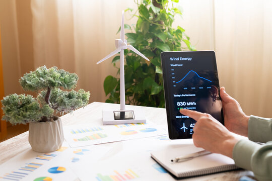 Person monitoring wind power output and energy statistics on a digital tablet screen, near a decorative wind turbine model and plant, highlighting renewable energy management
