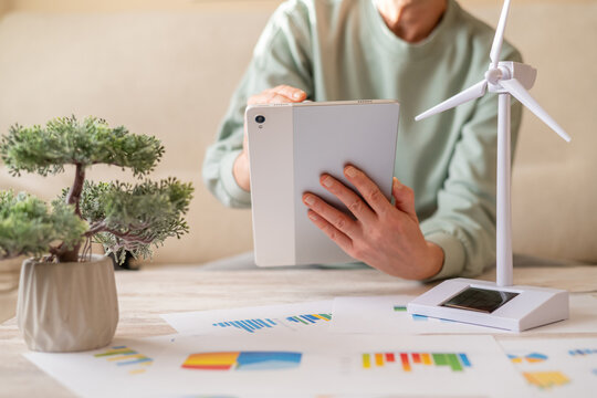 Person focusing on renewable energy project data, using a digital tablet next to a miniature wind turbine model and charts, representing green technology and future ecological solutions - Powered by Adobe