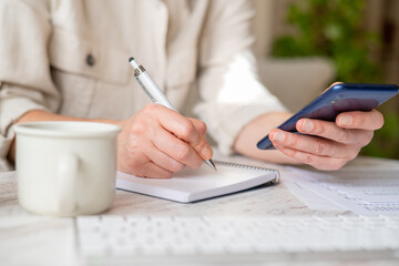 Fototapeta premium Woman managing personal finance, writing down expenses in a notebook while holding phone representing online banking or financial applications, focusing on home economy tasks