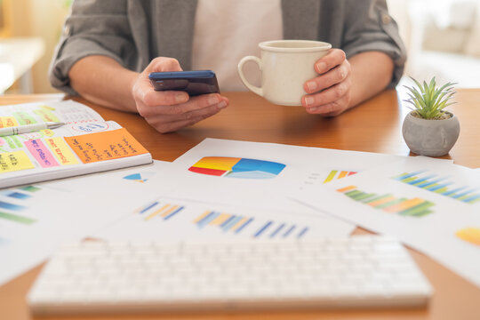 Person holding a smartphone and a coffee cup, analyzing finance data and business charts, planning work schedules, and managing personal economy from a home office desk - Powered by Adobe