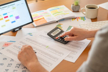 Person calculating finances at a desk with calculator, pen, financial documents, charts and tablet calendar, managing budgeting, expenses, taxes and planning for business or personal goals