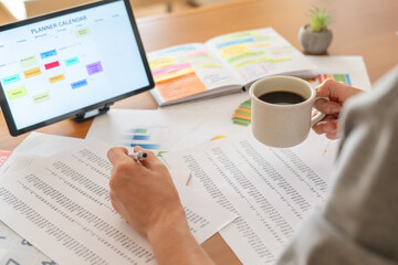 Hands holding a mug and pen over financial documents, tablet planner and open notebook on desk,...
