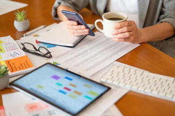 Woman reviewing budgets and financial reports at a home office desk, using smartphone and tablet while holding a coffee mug, calculating expenses and planning finances