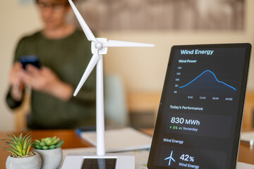 Person in blurred background using smartphone while tablet in foreground shows wind energy monitoring dashboard beside wind turbine model and potted plants, eco tech concept
