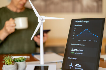 Person reading wind power generation data on a digital tablet with a desktop wind turbine model, symbolizing sustainable energy management and green technology