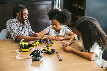 Teacher guides two girls building a robotics kit at a classroom table, assembling circuits and tools. Hands-on STEM learning and teamwork in a creative workshop environment.