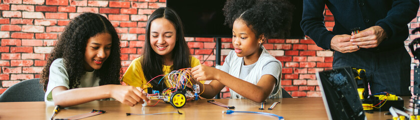 Three girls collaborate in classroom lab, assembling wheeled robot with wires. Guided by teacher,...