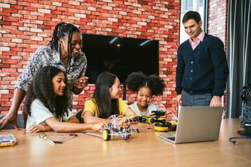 Diverse students and mentors brainstorm in classroom lab, coding and testing wheeled robot around laptop. Girls and boys collaborate with teacher, using parts and tools in hands-on STEM activity.