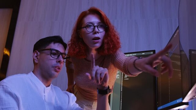 Two developers in glasses collaborate at glowing monitors with code in dim office. Late-night teamwork blends programming, database, strategy, planning, research, leadership, and consulting. 4k video